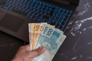 brazilian-money-banknotes-hand-with-blurred-background-laptop-computer-300x200